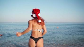 mujer sexy en traje de baño y sombrero de santa claus corre hacia el mar. la chica delgada se divierte mientras celebra el año nuevo en un país cálido cerca del océano. camara lenta.