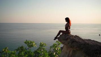 joven mujer delgada en traje de baño se sienta en una roca contra el telón de fondo de una puesta de sol y un paisaje marino. dama sexy en bikini se relaja cerca del mar. Hora de verano. vacaciones y viajes. video