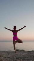 mujer fitness atlética en camiseta rosa y pantalones cortos haciendo yoga en el fondo del mar y las rocas al atardecer. una chica delgada realiza una pose espiritual y hace ejercicio al aire libre. libertad y motivación. vídeos verticales video