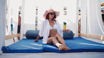 Slender woman in a hat sits relaxing and enjoys sunbathing in a gazebo on the sea beach on a bright sunny day. Summer vacation. video