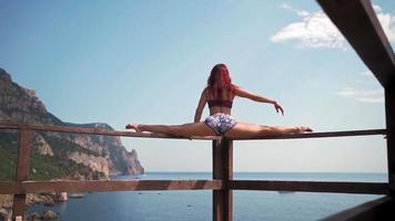 une fille athlétique est assise dans un grand écart sur fond d'un magnifique paysage de mer et de rochers. concept de voyage et de liberté. femme athlétique avec un étirement cool. ralenti. video