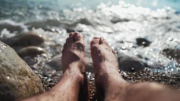 close-up nas pernas de um homem irreconhecível relaxando na praia do mar em um dia ensolarado de verão. conceito de férias e viagens. da primeira pessoa. pov video