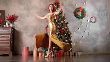 una hermosa mujer con un vestido de noche dorado gira y baila cerca de un árbol de navidad decorado con juguetes de año nuevo. Actitud festiva. material de archivo 4k de alta calidad