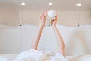 Young woman with a coffee mug in bed with white linens. Minimal happy morning photo