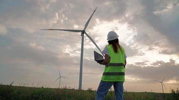 Woman Ecology Specialist in Uniform Checking with Laptop Correct Operation and Efficiency Windmills on Sunset. Female Engineer Monitoring System Performance and Making Notes in Program. video