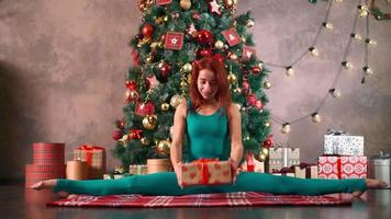 een atletisch slank vrouw in een turkoois jumpsuit zit in een splitst en opent een nieuw jaar geschenk tegen de achtergrond van een Kerstmis boom. gezond levensstijl concept. uitrekken en yoga.