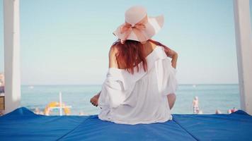 Schlanke Frau mit Hut sitzt entspannt und genießt das Sonnenbaden in einem Pavillon am Meeresstrand an einem sonnigen Tag. Sommerurlaub. video