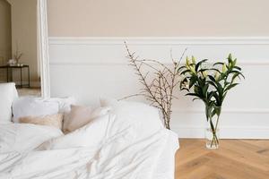 A bright cozy bed with pillows and lily flowers in a vase, tree branches. Scandinavian interior of the house photo