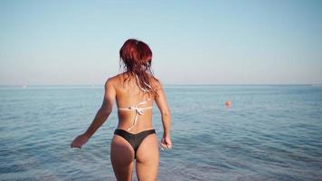 giovane contento donna con rosso capelli nel un' costume da bagno corre in il mare per un' nuotare. estate riposo. vacanza volta. lento movimento. video