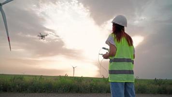 femme ingénieur en écologie en uniforme et casque tenant le joystick contrôlant le drone volant travaillant au moulin à vent sur fond de coucher de soleil magnifique. alternative à l'énergie électrique. video