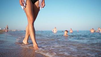 Sexy female legs are walking along the beach along the sea against the backdrop of bathing people. The woman walks barefoot splashing in the sea water. Summer vacation and relaxation. Slow motion. video