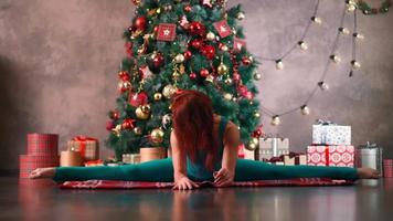 An athletic slim woman in a turquoise overalls practices yoga and stretching against the background of a Christmas tree. Weight loss after the New Year holidays. Healthy lifestyle concept. video