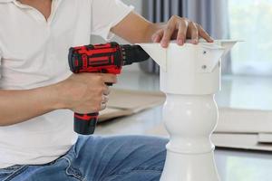 man assembling white table furniture at home using cordless screwdriver photo