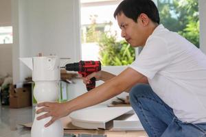 Asian man assembling white table furniture at home using cordless screwdriver photo