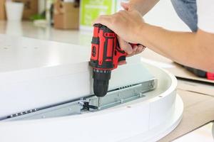 man assembling white table furniture at home using cordless screwdriver photo