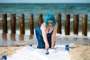 talentosa artista de performance de mujer de pelo azul untada con pinturas de gouache en un gran lienzo en la playa foto