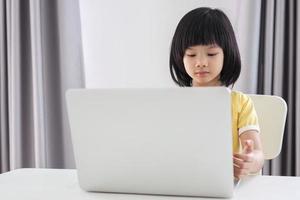 una pequeña estudiante asiática estudia en línea usando una computadora portátil en casa foto