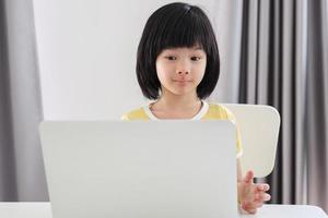 una pequeña estudiante asiática estudia en línea usando una computadora portátil en casa foto