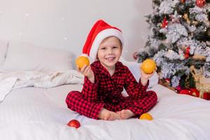 happy little boy in pajamas and Santa hat at home in bed eating tangerines for Christmas photo