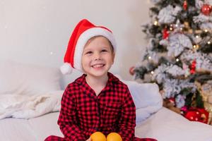 niño feliz en pijama y sombrero de santa en casa en la cama comiendo mandarinas para navidad foto