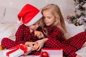 dos niños felices en pijama para navidad están clasificando regalos y jugando con juguetes navideños. foto de alta calidad