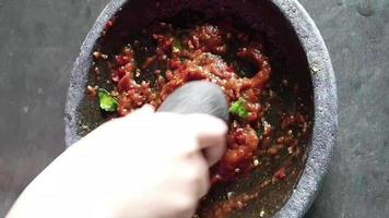 der Prozess der Herstellung einer Chilisauce. Eine Hand pulverisiert Zwiebeln und Chilis in einem Stößel auf einem Steinmörser, um eine Chilisauce herzustellen. video