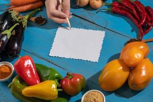 Vegetables are laid out around a sheet of paper and a pencil. Empty space for text. Vegetables, empty blank for recipe on a blue background. photo