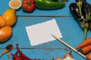 Vegetables are laid out around a sheet of paper and a pencil. Empty space for text. Vegetables, empty blank for recipe on a blue background. photo