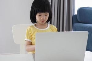 una pequeña estudiante asiática estudia en línea usando una computadora portátil en casa foto