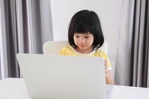 una pequeña estudiante asiática estudia en línea usando una computadora portátil en casa foto