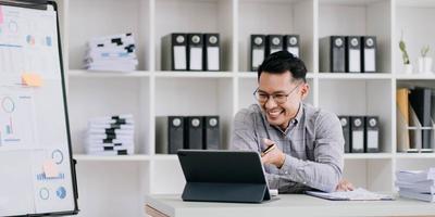 Portrait of a young Asian businessman smiling while using a laptop and writing down notes while sitting at his desk in a modern office photo