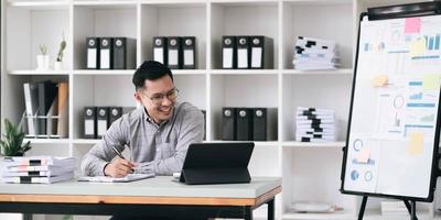 Portrait of a young Asian businessman smiling while using a laptop and writing down notes while sitting at his desk in a modern office photo