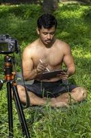 Young man practicing yoga asana, balance, meditating while standing on one leg on sports mat on green grass in park. Using tablet for online class. photo