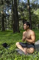 Young man practicing yoga asana, balance, meditating while standing on one leg on sports mat on green grass in park. Using tablet for online class. photo
