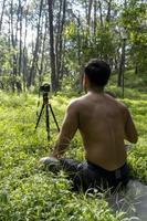 Young man practicing yoga asana, balance, meditating while standing on one leg on sports mat on green grass in park. Using tablet for online class. photo