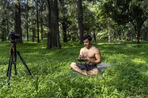 Young man practicing yoga asana, balance, meditating while standing on one leg on sports mat on green grass in park. Using tablet for online class. photo