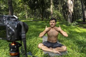 Young man practicing yoga asana, balance, meditating while standing on one leg on sports mat on green grass in park. Using tablet for online class. photo