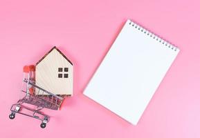 flat layout of wooden house model in shopping trolley and  blank page notebook  on pink  background with copy space, home purchase concept. photo