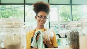 A young Black female shopkeeper cleans glass jars of natural organic products in reusable containers at a refill store, zero waste, a plastic-free grocery shop, and an eco-friendly retail business. video