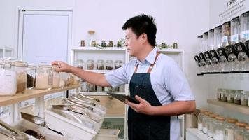 Asian male shopkeeper checks stock of organic products at ingredients shelves in refill store, zero-waste and sustainable shop, Thai texts on the wall mean fill a container with any bulk items video