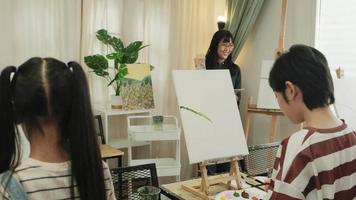 A female Asian teacher teaches and demonstrates to the children on acrylic color picture painting on canvas in art classroom, creatively learning with skill at the elementary school studio education. video