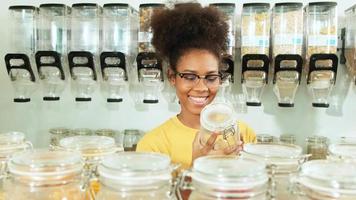Young African American female customer chooses and shop for organic products in refill store, zero-waste grocery, plastic-free, reusable containers, and an environment-friendly sustainable lifestyle.