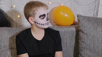 retrato de close-up de um menino com maquiagem no rosto na forma de um esqueleto para o halloween. criança está pronta para o feriado de doces ou travessuras. video