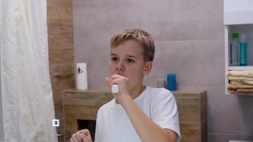 een vrolijk tiener borstels zijn tanden met een tandenborstel in de ochtend- in de badkamer en zingt. glimlachen knap schooljongen is genieten van ochtend- routines. video