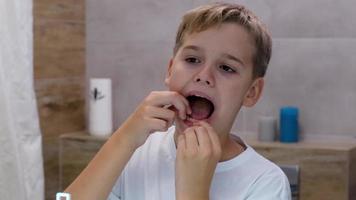 feche o reflexo do espelho de tiro na cabeça de um menino feliz passando fio dental nos dentes. estudante bonito desfrutando de sua rotina de higiene oral matinal sozinho no banheiro. video
