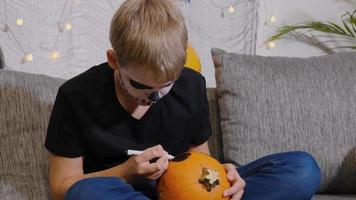 Ein Junge mit einem bemalten Skelett im Gesicht malt einen Kürbis für Halloween. Kind ist bereit für den Süßes oder Saures Urlaub. video