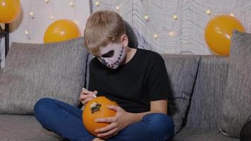 un niño con maquillaje de esqueleto pinta una calabaza para halloween mientras está sentado en el sofá de casa. el niño está listo para las vacaciones de truco o trato. video