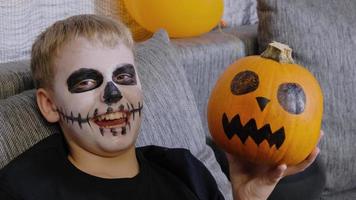 A scary child with a make-up in the form of a skeleton and with a pumpkin in his hands laughs looking at the camera. The child celebrates Halloween video
