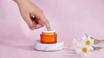 Skincare product in orange container on marble platform with flower beside video