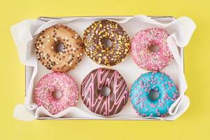 Different types of colorful donats decorated sprinkles and icing in box on a yellow background, top view photo